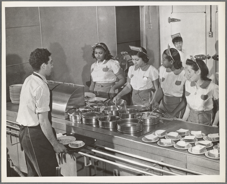 [A man standing with a tray of food at a food service counter and looking at four women who are standing behind the counter and spooning food onto plates.],Food preparation and service., Digital ID 1260375, New York Public Library