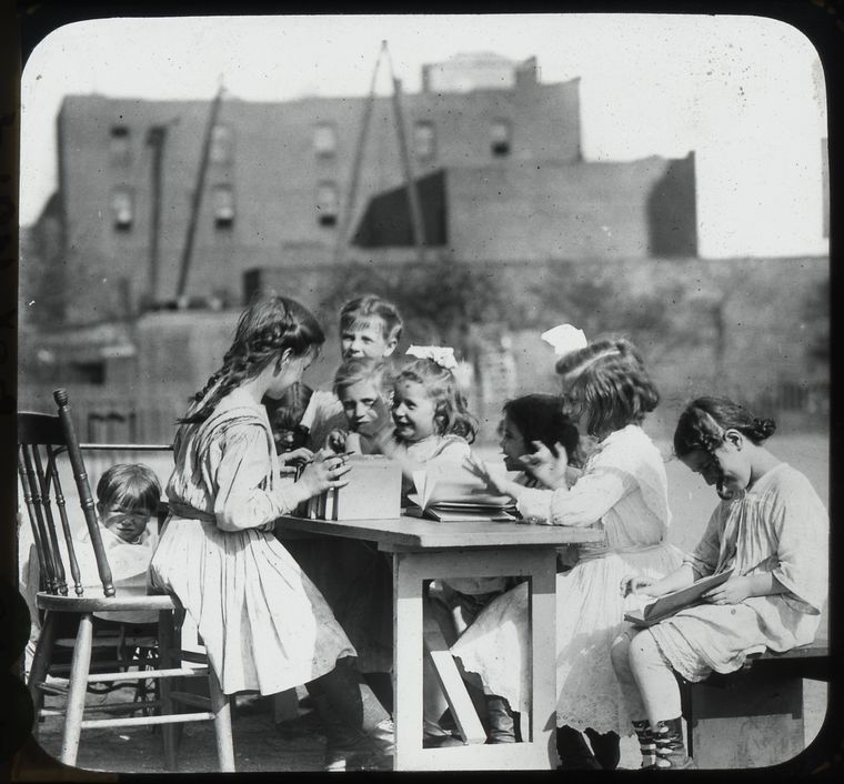  older girl with books on one side of table in playground, other children on the other side. , Digital ID 434224, New York Public Library