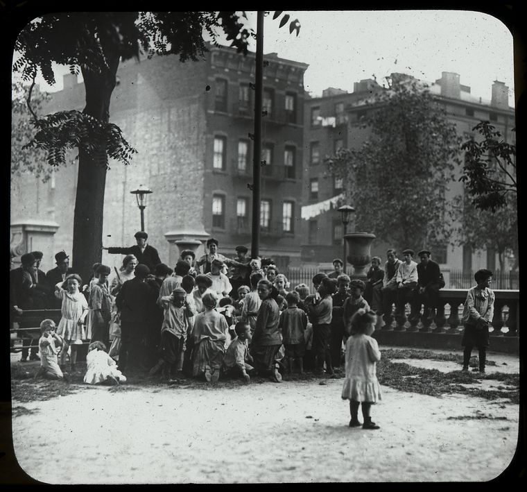  men listen along with children to story in the park, neighborhood buildings visible beyond, ca. 1910s., Digital ID 465199, New York Public Library