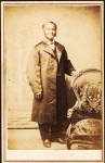 Studio portrait of unidentified man, standing, wearing long coat