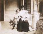 Postcard of an unidentified woman, probably a nanny, sitting on a porch with two white children.