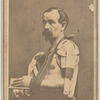 Shirtless seated man displays left arm in splint, resting in sling of striped cloth