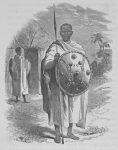 Male standing with spear and shield in hand, another man with spear and shield standing next to hut in background