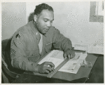 African American First Lieutenant Seth Finley seated at a desk and holding a ruler over a stack of papers, Fort Lewis, Washington