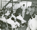 African American chaplain standing at the podium and conducting the religious service on deck of a U. S. Coast Guard vessel, South Pacific