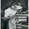 African American woman holding a perforated metallic box frame up to a piece of metalworking equipment