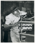 African American woman holding a perforated metallic box frame up to a piece of metalworking equipment
