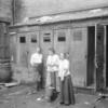 Two women & man in front of outhouses; one woman getting water