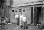 Two women & man in front of outhouses; one woman getting water