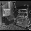 Kitchen with stove (having two irons and kettle and hot water tank above it), cupboard (filled with dishes), and table (covered with figured cloth)