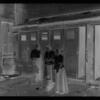 Two women & man in front of outhouses; one woman getting water
