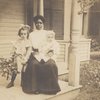 Postcard of an unidentified woman, probably a nanny, sitting on a porch with two white children.