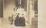 Postcard of an unidentified woman, probably a nanny, sitting on a porch with two white children.
