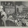 An audience of service men and hostesses watching George Jessel and an unidentified woman on stage.