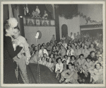 An audience of service men and hostesses watching George Jessel and an unidentified woman on stage.