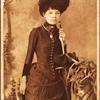 Standing woman, wearing dress with bustle, holding purse at side and umbrella resting on pedestal.