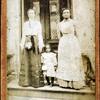 Two women and a child standing on step in front of a doorway of a house.