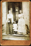 Two women and a child standing on step in front of a doorway of a house.