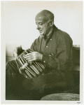 Henry Cowell trying out one of the five tablas for the tablatarang (= circle of tuned drums) presented to him for his Symphony No. 13 (Madras) by the Music Academy of Madras, India.  NYC, 1959