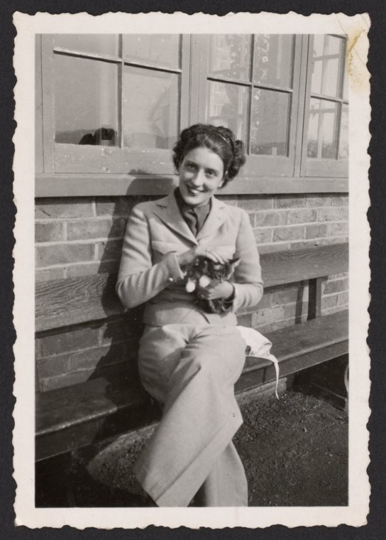 Portrait photograph of woman in winter coat, sitting on a bench, petting a cat that is sitting on her lap.