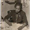 Claudia Jones seated at her desk reviewing a copy of the West Indian Gazette, London