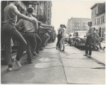 Jerome Robbins, crew, dancers: Exterior, city sidewalk: Jerome Robbins conducting warm-up class, row of male dancers in street clothes using basement railings for barre