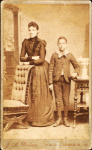 Unidentified standing figures: woman, her folded arms leaning on back of chair, and young man, probably her son, left hand on book.