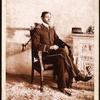 Uniformed man seated, on the mantlepiece is his cap, embroidered with the name Miller Edison Phonograph Co. 