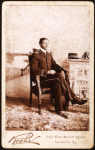 Uniformed man seated, on the mantlepiece is his cap, embroidered with the name Miller Edison Phonograph Co. 