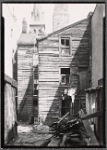 Debris strewn yard; wooden tenement; church steeple: Manhattan