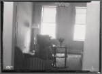 Woman seated in living room; newspaper with Rockefeller death headline