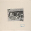 Several people in front of thatch roof structure, one, apparently kneeling or seated on the ground, is wearing a poncho which covers him from the neck down