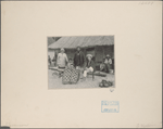Several people in front of thatch roof structure, one, apparently kneeling or seated on the ground, is wearing a poncho which covers him from the neck down