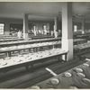 Large dining hall, empty except for about ten members of the dining hall staff. The place settings consist of a worn porcelain-enameled plate, a fork and knife.