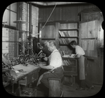 Enos Company : man works at workbench as another chooses books from shelves in corner, July 29, 1910