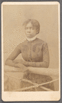 Unidentified young woman wearing gloves, leaning on prop fence.