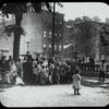 Work with schools, Hudson Park Branch : men listen along with children to story in the park, neighborhood buildings visible beyond, ca. 1910s