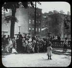 Work with schools, Hudson Park Branch : men listen along with children to story in the park, neighborhood buildings visible beyond, ca. 1910s