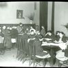 Work with schools : waiting their turn : children lined up at librarian's desk, others work at tables, ca. 1910s