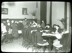 Work with schools : waiting their turn : children lined up at librarian's desk, others work at tables, ca. 1910s
