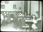 Work with schools : waiting their turn : children lined up at librarian's desk, others work at tables, ca. 1910s