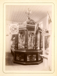 [A glass enclosed hexagonal cabinet filled with flowers nad glass jars.   A double headed eagle on the top of the cabinet.]