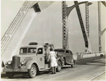 Model T 23 on a bridge with a car being pulled up to tow; a person wearing trench coat and fedora hat oversees