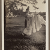 Edith James Long outdoors, dressed in Chinese costume with long scarves holding pole and basket.