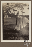 Edith James Long outdoors, dressed in Chinese costume with long scarves holding pole and basket.