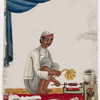 Seated male food vendor roasting meat on spit,  fanning flames