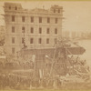 The obelisk "Cleopatra's Needle" in a horizontal position with men standing on it as they secure it for transport, city waterfront visible