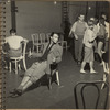 James Mitchell seated, wearing rolled up trousers, suit jacket and scarf, holding a sword and a cane during a rehearsal of Brigadoon