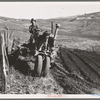 Young farmer plowing while other co-op members work in the sawmill. The tractor does work for five member families. Ola self-help sawmill co-op. Gem County, Idaho