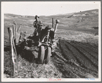 Young farmer plowing while other co-op members work in the sawmill. The tractor does work for five member families. Ola self-help sawmill co-op. Gem County, Idaho